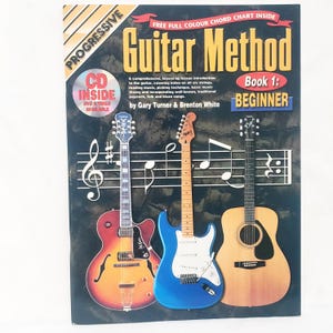 May include: A guitar method book titled "Guitar Method Book 1: Beginner" with three guitars. The cover features a red, blue, and natural wood guitar. The book includes a CD and DVD, and a free full-color chord chart.