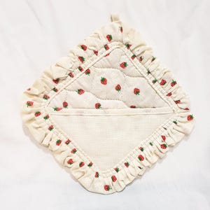 May include: Cream-coloured quilted pot holder with a strawberry pattern. The pot holder has a ruffled edge and a loop for hanging. The centre features a quilted design with small red strawberries. The overall design is a charming, country-style aesthetic.