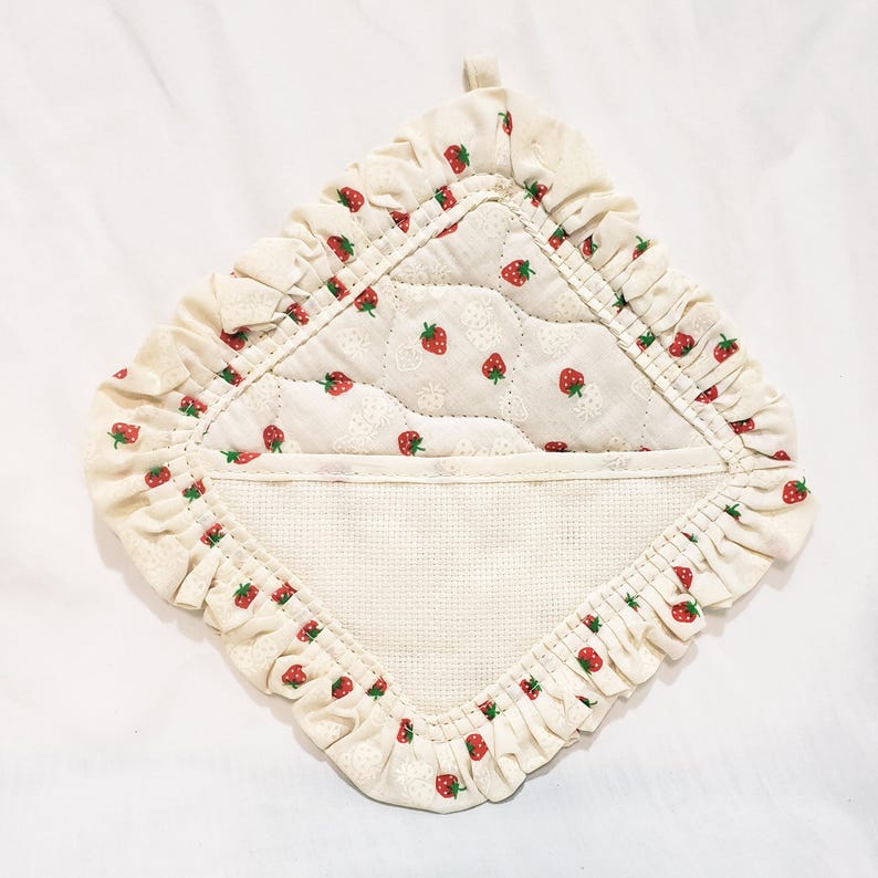 May include: Cream-coloured quilted pot holder with a strawberry pattern. The pot holder has a ruffled edge and a loop for hanging. The centre features a quilted design with small red strawberries. The overall design is a charming, country-style aesthetic.