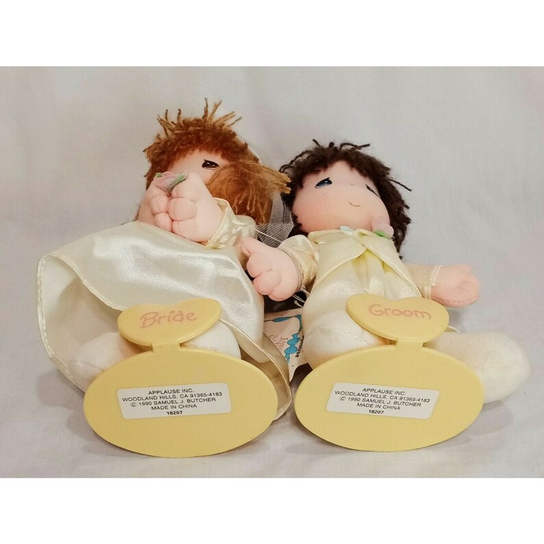 May include: A pair of plush bride and groom dolls with embroidered details. The bride doll is wearing a white dress with a veil and holding a bouquet of flowers. The groom doll is wearing a white suit with a bow tie. Both dolls have a heart-shaped tag on their feet that says "Bride" and "Groom". The dolls are made by Applause Inc. and were made in China.
