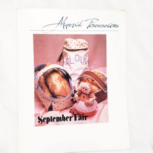 May include: A vintage craft pattern booklet titled "Material Possessions: September Fair". The cover features a pink background with a doll, a bread basket, and a flour sack.