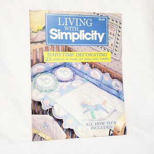 Könnte beinhalten: Ein Buchcover mit einem blau-weißen Kinderbett und einer Steppdecke mit einem Schaukelpferd-Motiv. Der Buchtitel lautet "Living with Simplicity: Babytime Decorating 23 Projekte zum Selbermachen für Baby und Kleinkind. Alle Anleitungen enthalten."