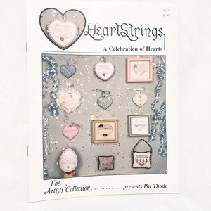 May include: A magazine cover titled "HeartStrings: A Celebration of Hearts" featuring various heart-shaped cross-stitch designs. The designs include framed pieces and hanging ornaments in soft pastel colors, with text and decorative elements.