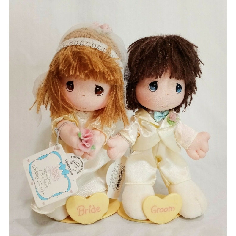 May include: A bride and groom Precious Moments doll set. The bride doll is wearing a white dress and veil, holding a pink rose. The groom doll is wearing a white tuxedo with a blue bow tie. Both dolls are standing on heart-shaped bases that say "Bride" and "Groom".