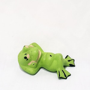 Könnte beinhalten: Eine grüne Keramik-Froschfigur mit schwarzen Augen und geschlossenem Mund, die auf dem Rücken liegt und die Beine ausgestreckt hat.