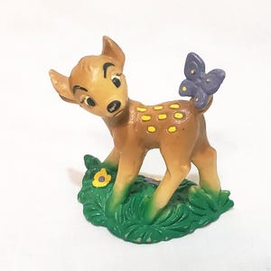 May include: A brown deer figurine with yellow spots and a purple butterfly on its back. The deer is standing on a green base with a yellow flower. The figurine has black eyes and is looking to the left.