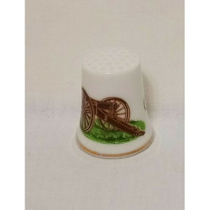 May include: White porcelain thimble with a brown and green image of a cannon on a grassy field.