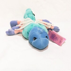 May include: A plush toy in the shape of a blue and teal creature with white accents and pink and purple limbs. The toy has a tag that reads "Precious Moments Tender Toys". The toy is lying on a white surface.