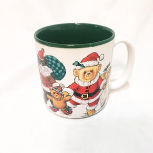 Op de afbeelding: Witte keramische mok met een groene binnenrand en een wit handvat. De mok heeft een feestelijk ontwerp van teddyberen gekleed in kerstman outfits, die cadeaus dragen en kerstmutsen dragen. Een mok met een vakantiethema.