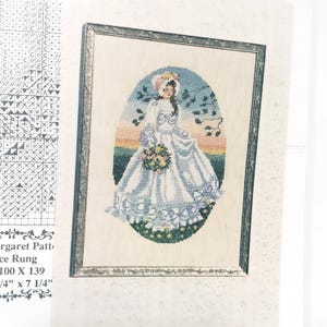May include: A cross-stitch pattern featuring a person in a long white gown and bonnet holding a bouquet of flowers. The design is framed in an oval against a sunset background with green foliage. The pattern includes a grid and text reading Margaret Pattern, 100 X 139, 7 1/4" x 7 1/4".