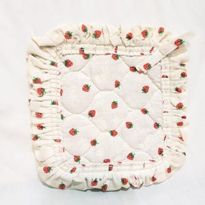 May include: A square, quilted pot holder featuring a white background adorned with a repeating pattern of red strawberries. The edges are trimmed with a ruffled fabric, also decorated with strawberries. A small fabric loop is attached.