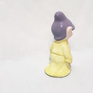 May include: A small ceramic figurine of a dwarf wearing a yellow robe and a purple hat. The dwarf is standing with his arms at his sides and is looking to the right.