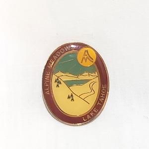 Op de afbeelding: Ovale speld met een landschapsontwerp. De speld toont een meer, bergen en een weg, met de tekst "ALPINE MEADOW LAKE TAHOE". De speld heeft een gouden rand en een gouden letter "A" in een cirkel.