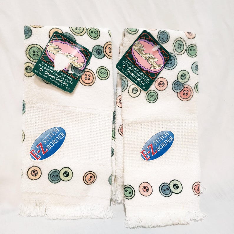 May include: Two white hand towels with a button pattern in pastel colors. Each towel has a fringed edge and a blue oval sticker that reads "E-Z STITCH BORDER." Tags with the text "Charles Craft" are attached.