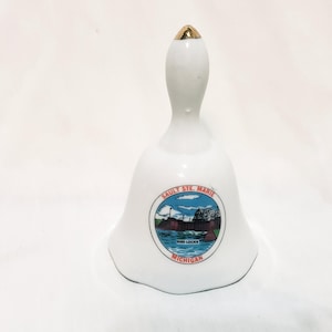 May include: White ceramic bell with a gold-colored top. The bell features a circular graphic with the text "SAULT STE. MARIE" and "500 LOCKS MICHIGAN". The graphic depicts a boat and buildings.