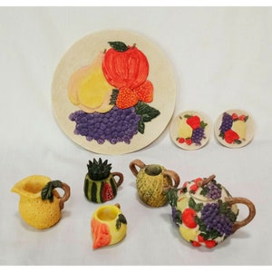 May include: A set of miniature teacups and teapot decorated with fruit and grape designs. The set includes a teapot, a creamer, a sugar bowl, and two saucers. The teacups and teapot are made of ceramic and have a white base with colorful fruit and grape designs.