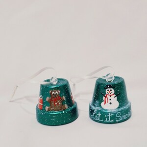 May include: Two green Christmas ornaments with white glitter. One ornament features a snowman with the text "Let it Snow". The other ornament features a teddy bear holding a gift.