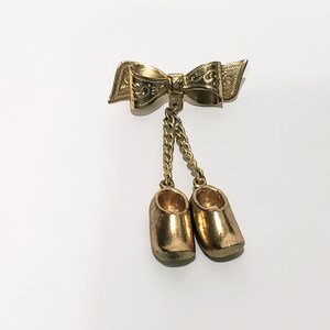 May include: Gold-toned brooch featuring a bow with intricate details and two dangling charms shaped like wooden shoes.