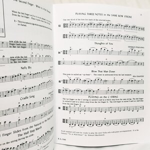 May include: An open music instruction book for viola featuring printed musical notation, staff lines, and instructional text including titles like Playing Three Notes in the Same Bow Stroke, Thoughts of You, Blow That Man Down, and Playing on the C String.
