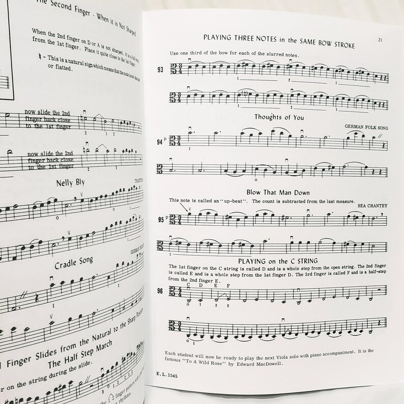 May include: An open music instruction book for viola featuring printed musical notation, staff lines, and instructional text including titles like Playing Three Notes in the Same Bow Stroke, Thoughts of You, Blow That Man Down, and Playing on the C String.