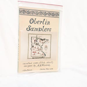 Puede incluir: Un patrón de punto de cruz contado titulado Oberlin Samplers que presenta un mapa de Minnesota con varios puntos de referencia e iconos. El patrón de papel está encerrado en una funda de plástico transparente e incluye el texto designed by M.M. Waldock.