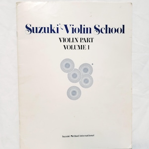 May include: A white book cover with the title "Suzuki Violin School" in blue text. The subtitle is "Violin Part Volume I" in smaller blue text. There is a blue and white abstract design in the center of the cover. The bottom of the cover reads "Suzuki Method International" in blue text.