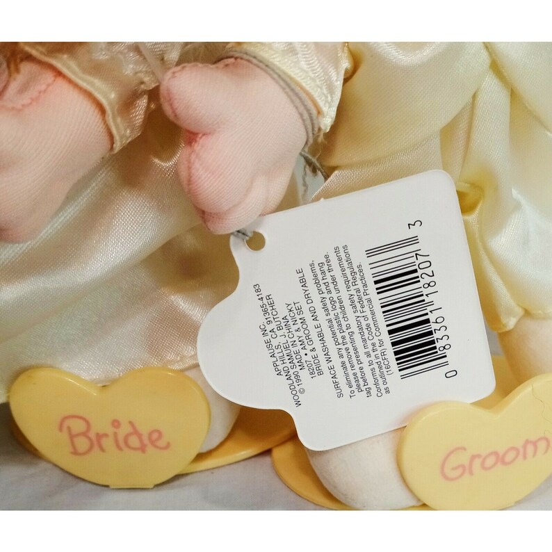May include: A bride and groom doll set with yellow heart-shaped bases. The dolls are dressed in white satin and have pink faces. The tag reads "Bride & Groom Set" and includes information about the manufacturer and care instructions.