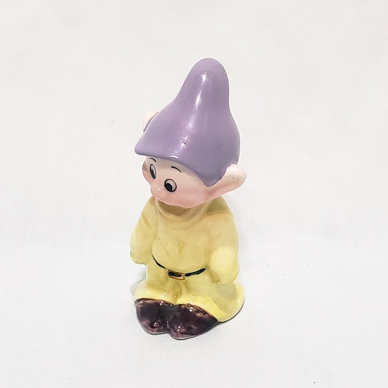 May include: A small ceramic figurine of a dwarf wearing a purple hat and yellow clothing. The dwarf has a friendly expression and is standing with his feet together.