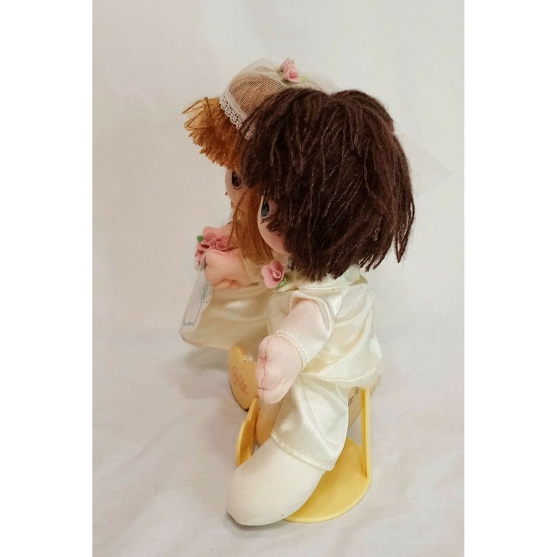 May include: A pair of plush bride and groom dolls dressed in white satin with lace trim. The bride has brown hair and a veil, while the groom has brown hair. They are holding hands and sitting on a yellow base.