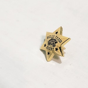 May include: A gold-colored star-shaped pin with the words "Wells Fargo Agent" and a stagecoach design in black. The pin has a classic, vintage aesthetic.