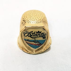 Op de afbeelding: Een goudkleurige vingerhoed met een schildvormig embleem. Het embleem toont het woord "Atlantic" boven een illustratie van een schip op blauw water. De vingerhoed is een naai-instrument en de tekst "Star/Ship" staat boven het woord "Atlantic".