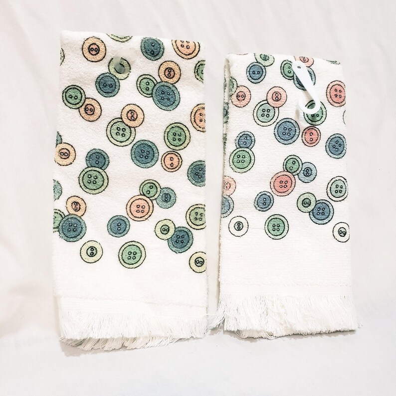 May include: Two white cotton hand towels with a repeating pattern of colorful buttons in shades of blue, green, orange, and pink. The towels have a fringed edge at the bottom. A white plastic clip is attached to one of the towels.