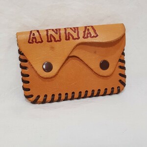 Puede incluir: Una pequeña cartera de cuero de color marrón claro con un borde cosido de color marrón. La cartera tiene dos broches a presión y el nombre "ANNA" está grabado en rojo en la parte delantera.