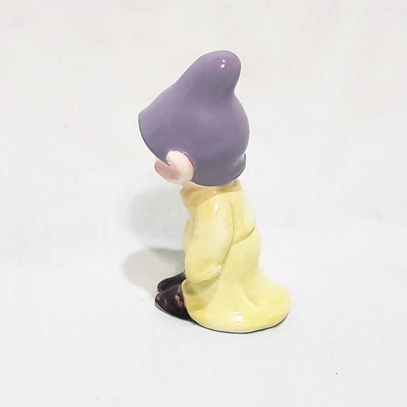 May include: A ceramic figurine of a dwarf from the movie Snow White and the Seven Dwarfs. The dwarf is wearing a purple hat and a yellow coat. He is looking down and to the left.