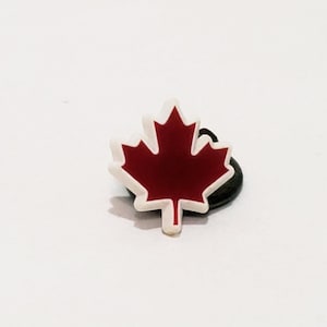 May include: A red maple leaf charm with a white outline. The charm is attached to a black pin.