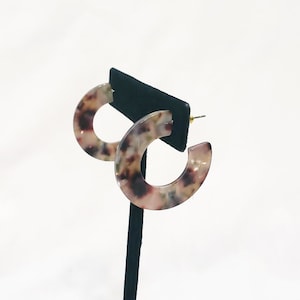 May include: A pair of hoop earrings with a tortoiseshell pattern in shades of brown, pink, and white. The earrings are displayed on a black earring stand.