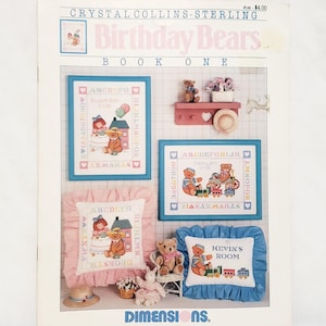 May include: A cross-stitch pattern booklet featuring a collection of birthday bear designs. The cover shows a pink pillow with a cross-stitch design of a girl and a bear having tea, a blue pillow with a cross-stitch design of two bears riding a train, and a framed cross-stitch design of a girl and a bear with balloons. The booklet is titled "Birthday Bears Book One" and is by Crystal Collins-Sterling.