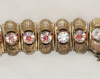 Puede incluir: Un brazalete dorado con una fila de piedras transparentes iridiscentes engastadas en engastes de metal ornamentados.