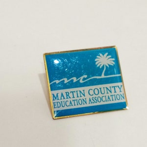 Può includere: Spilla quadrata con sfondo azzurro chiaro e bordi dorati. La spilla presenta una palma bianca e il testo "MARTIN COUNTY EDUCATION ASSOCIATION". Sotto, grafiche di onde bianche.