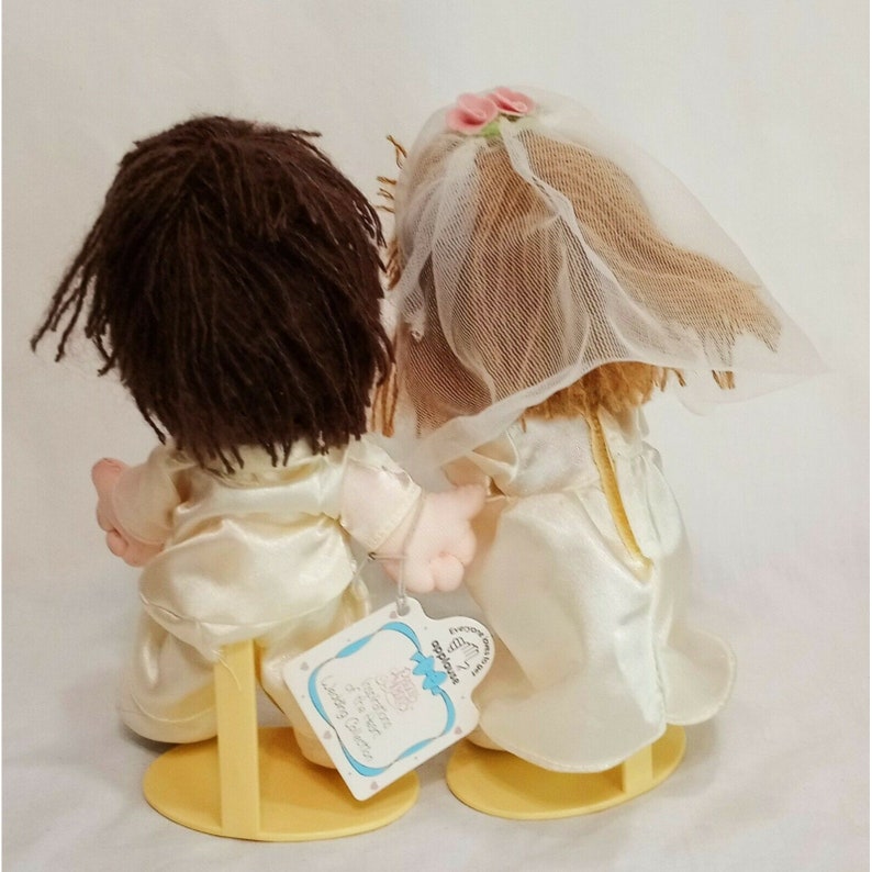 May include: A pair of plush wedding dolls, a bride and groom, wearing white satin dresses and suits. The bride has a white veil and pink flowers in her hair. The dolls are standing on yellow plastic stands. The tag reads "Forever Friends, Little Couple"