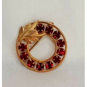 May include: A gold-toned brooch with a red rhinestone wreath and a gold leaf design.