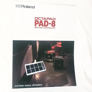 Könnte beinhalten: Ein weißes Roland Octapad Pad-8 MIDI-Pad-Controller mit acht schwarzen Pads, das auf einer Bühne gezeigt wird. Das Bild enthält das Roland-Logo und den Text "Electronic Musical Instruments."