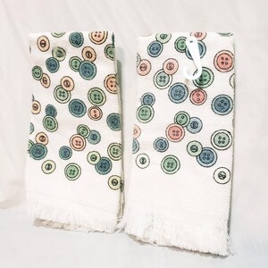 May include: Two white hand towels with a pattern of colorful buttons in shades of blue, green, orange, and pink. The towels have a fringed edge and a white plastic hook.