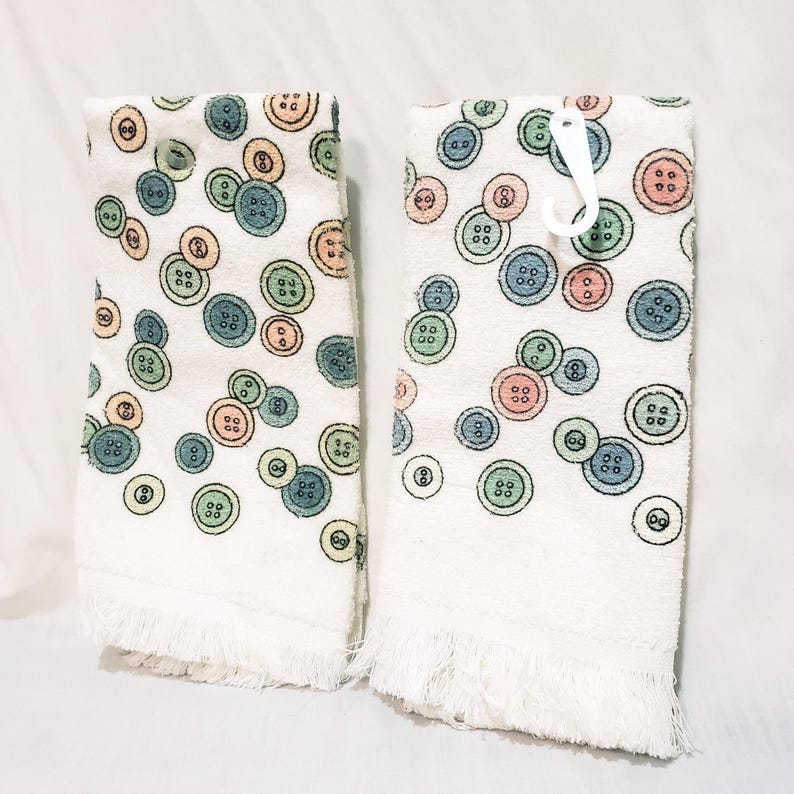 May include: Two white hand towels with a pattern of colorful buttons in shades of blue, green, orange, and pink. The towels have a fringed edge and a white plastic hook.