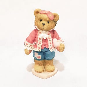 May include: A brown teddy bear figurine wearing a pink jacket with heart accents, blue shorts, and a pink eye mask. The bear holds a pink heart and stands on a heart-shaped base. The figurine has a vintage aesthetic.