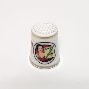 May include: White ceramic thimble with a gold trim and a Sen-Sen candy advertisement featuring a woman in a red dress. The advertisement includes the text "Sen-Sen 5c Sen-Sen".