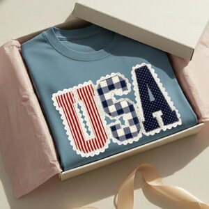 May include: A light blue crew neck sweatshirt with the word "USA" appliqued on the front. The letters are in red and white stripes, blue and white gingham, and navy blue with white polka dots, all with a white scalloped border. The sweatshirt is in a white gift box.