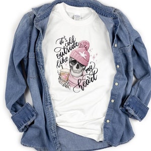 May include: White t-shirt with a graphic of a skull wearing a pink beanie and scarf, holding a coffee cup. The text reads "It's cold outside like my heart" in black script. A denim jacket is draped over the shirt.