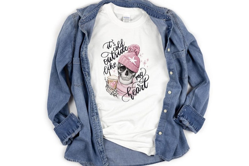 May include: White t-shirt with a graphic of a skull wearing a pink beanie and scarf, holding a coffee cup. The text reads "It's cold outside like my heart" in black script. A denim jacket is draped over the shirt.