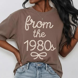May include: A brown t-shirt with the text "From the 1980s" in a pearl-like font. The shirt has short sleeves and a round neckline. The text is arranged in a decorative style with a bow at the bottom.
